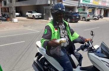 Pengemudi motor mengenakan jaket hijau dan helm hitam di jalan kota.